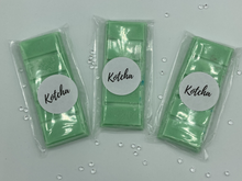 Load image into Gallery viewer, The picture is of a wax melt in the shape of a chocolate bar. The colour is green with silver biodegradable glitter wrapped in plastic with the brand name "Kotcha" on it. There are crystals surrounding the image and a white background.