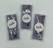 Load image into Gallery viewer, The picture is of a wax melt in the shape of a chocolate bar. The colour is purple with black biodegradable glitter wrapped in plastic with the brand name "Kotcha" on it. There are crystals surrounding the image and a white background.