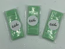 Load image into Gallery viewer, The picture is of a wax melt in the shape of a chocolate bar. The colour is green with silver biodegradable glitter wrapped in plastic with the brand name &quot;Kotcha&quot; on it. There are crystals surrounding the image and a white background.
