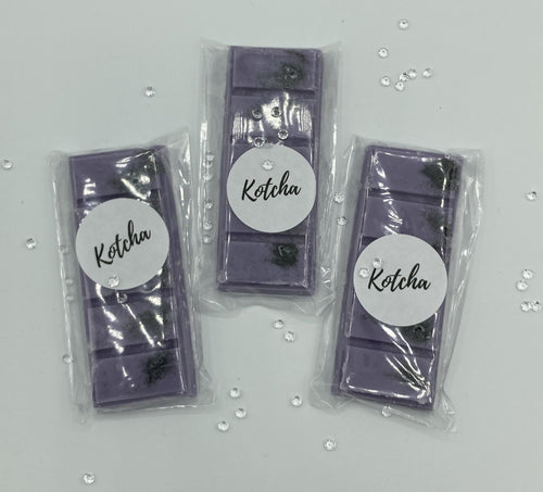 The picture is of a wax melt in the shape of a chocolate bar. The colour is purple with black biodegradable glitter wrapped in plastic with the brand name 