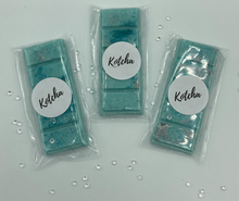 Load image into Gallery viewer, The picture is of a wax melt in the shape of a chocolate bar. The colour is green and blue (teal)  with silver biodegradable glitter wrapped in plastic with the brand name &quot;Kotcha&quot; on it. There are crystals surrounding the image and a white background.
